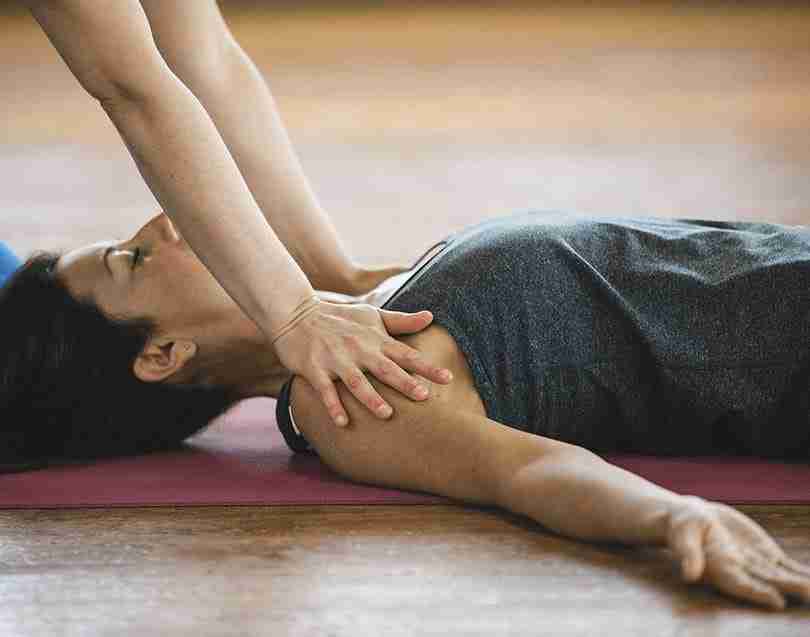 Chica en el suelo sobre mat de yoga siendo asistida por la profesora de yoga en una clase de yoga a domicilio